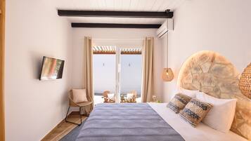Un dormitorio con cabecera de piedra, una cama con manta azul, una silla de mimbre y vistas al mar a través de un ventanal.