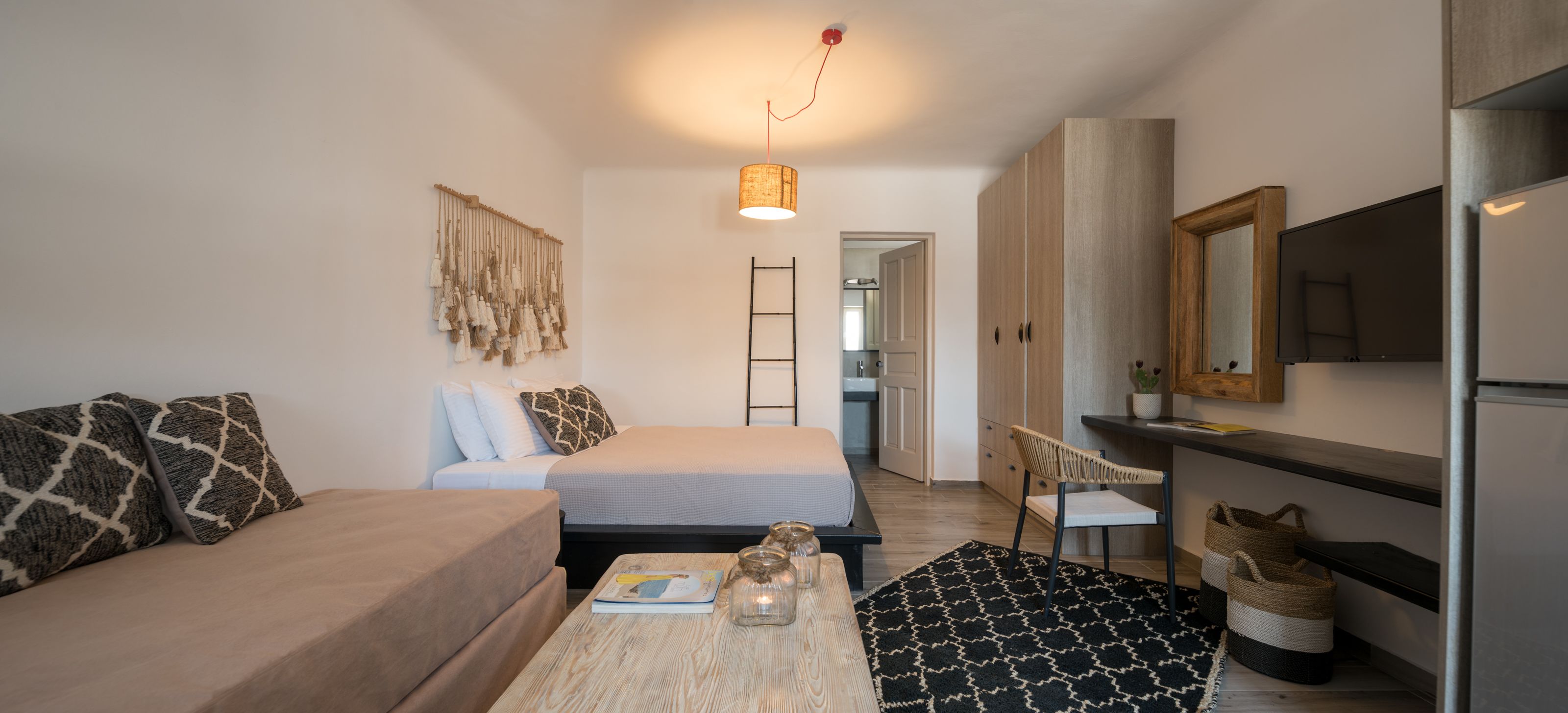 Ett litet rum med en säng, en soffa, ett litet bord med en tidning, en hängande lampa, en stol, en tv och ett kylskåp.
