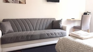 Un sofá gris con un reposabrazos blanco, un tapete estampado y una mesita con un paño blanco doblado.