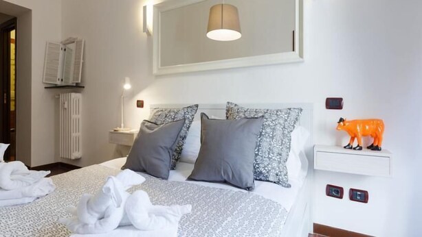 Habitación de hotel con cama, almohadas, mesita de noche, lámpara de pared, radiador y una pequeña figurina naranja.