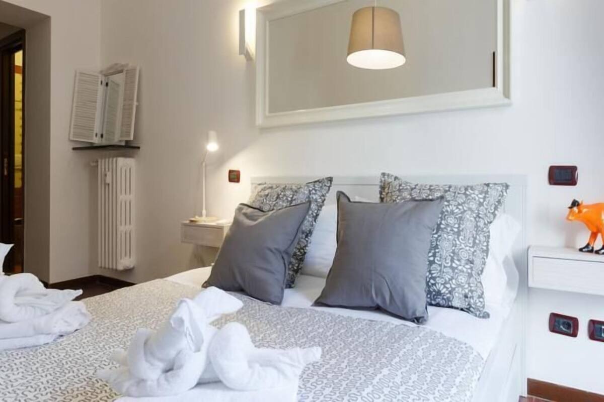 A hotel room with a bed, pillows, a nightstand, a wall-mounted lamp, a radiator, and a small orange figurine.
