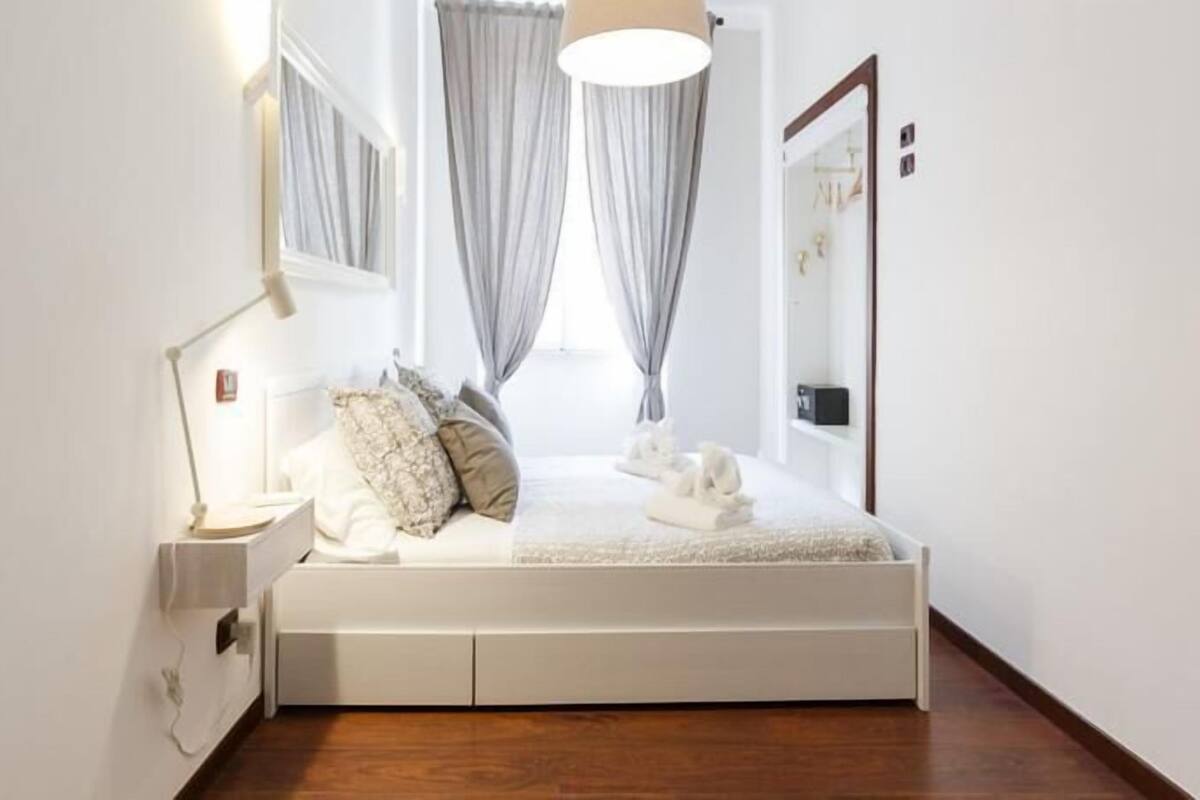 A bedroom with a white bed, a bedside lamp, and a window with curtains.