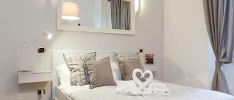 Una cama bien hecha con una toalla dispuesta en forma de corazón, una mesita de noche con lámpara y un espejo sobre la cama.