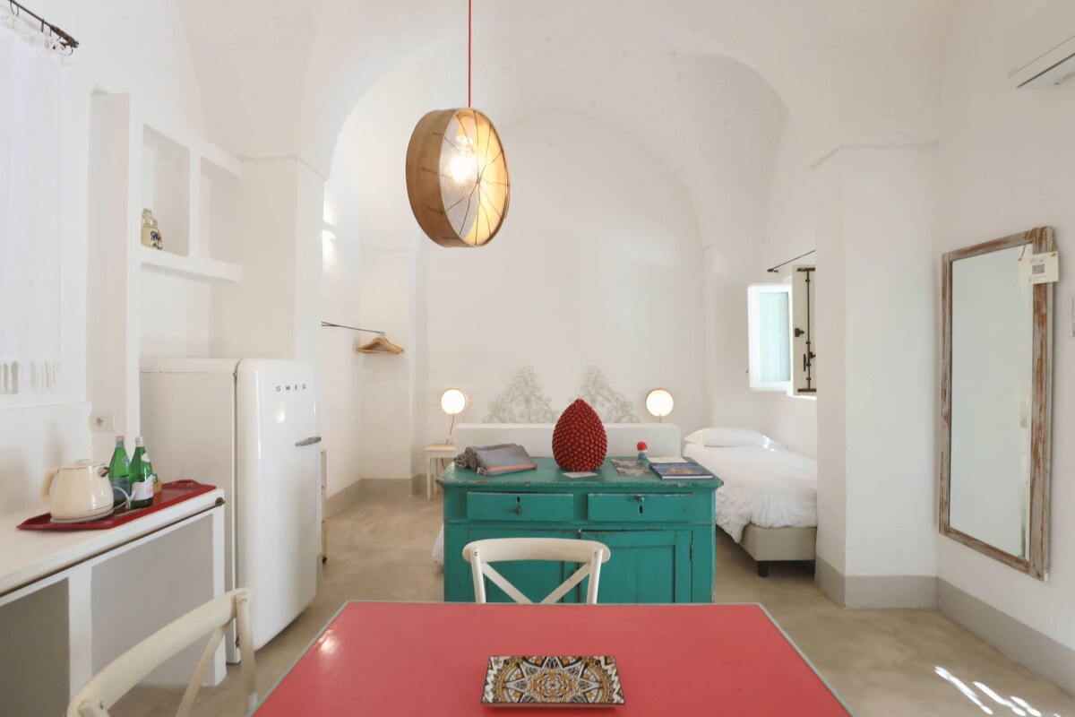 Una habitación compacta con kitchenette, una cama, un aparador azul verdoso y una lámpara colgante.