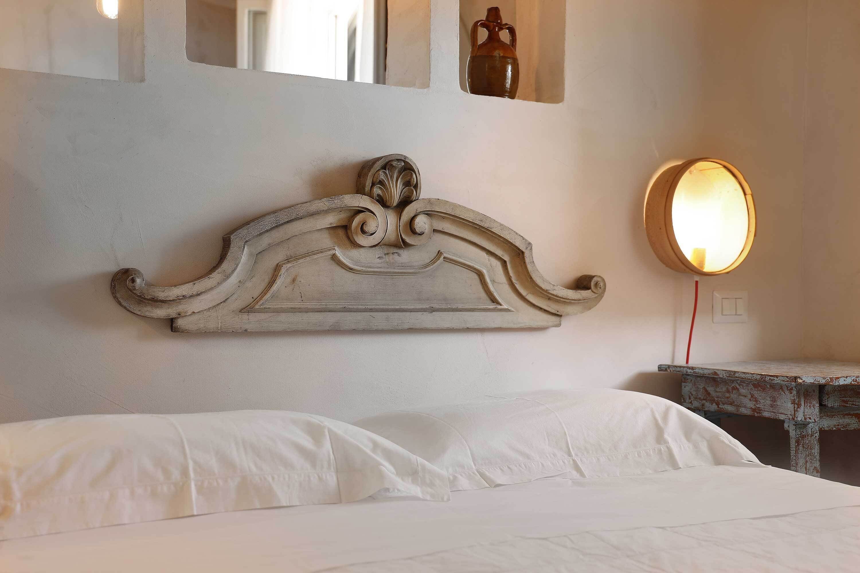 A bed with white linens, a decorative headboard, a small bedside table, and a wall-mounted mirror with a light.