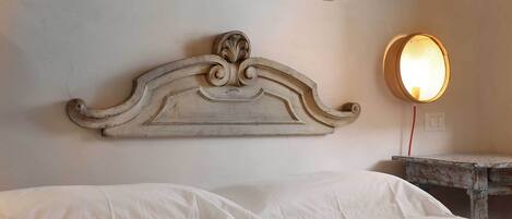 A bed with white linens, a decorative headboard, a small bedside table, and a wall-mounted mirror with a light.