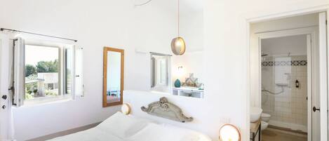 Quarto com cama, mesa de cabeceira, espelho, janela e banheiro visível através de uma porta aberta.