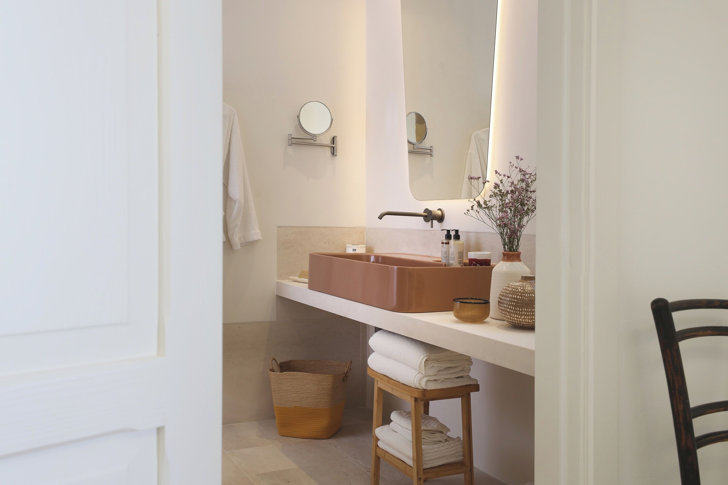Un baño con un lavamanos blanco, un espejo, un perchero para toallas y un taburete de madera pequeño.