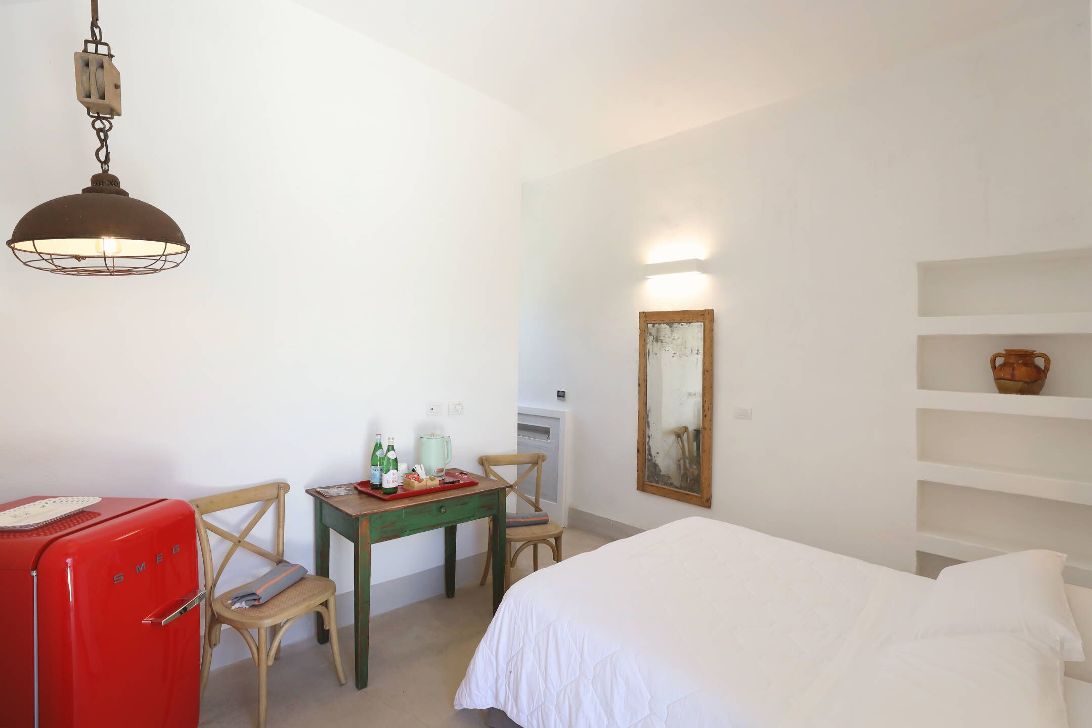 Una habitación con una cama, un escritorio con silla, un refrigerador rojo de estilo vintage, una lámpara de pared y una estantería con un florero.