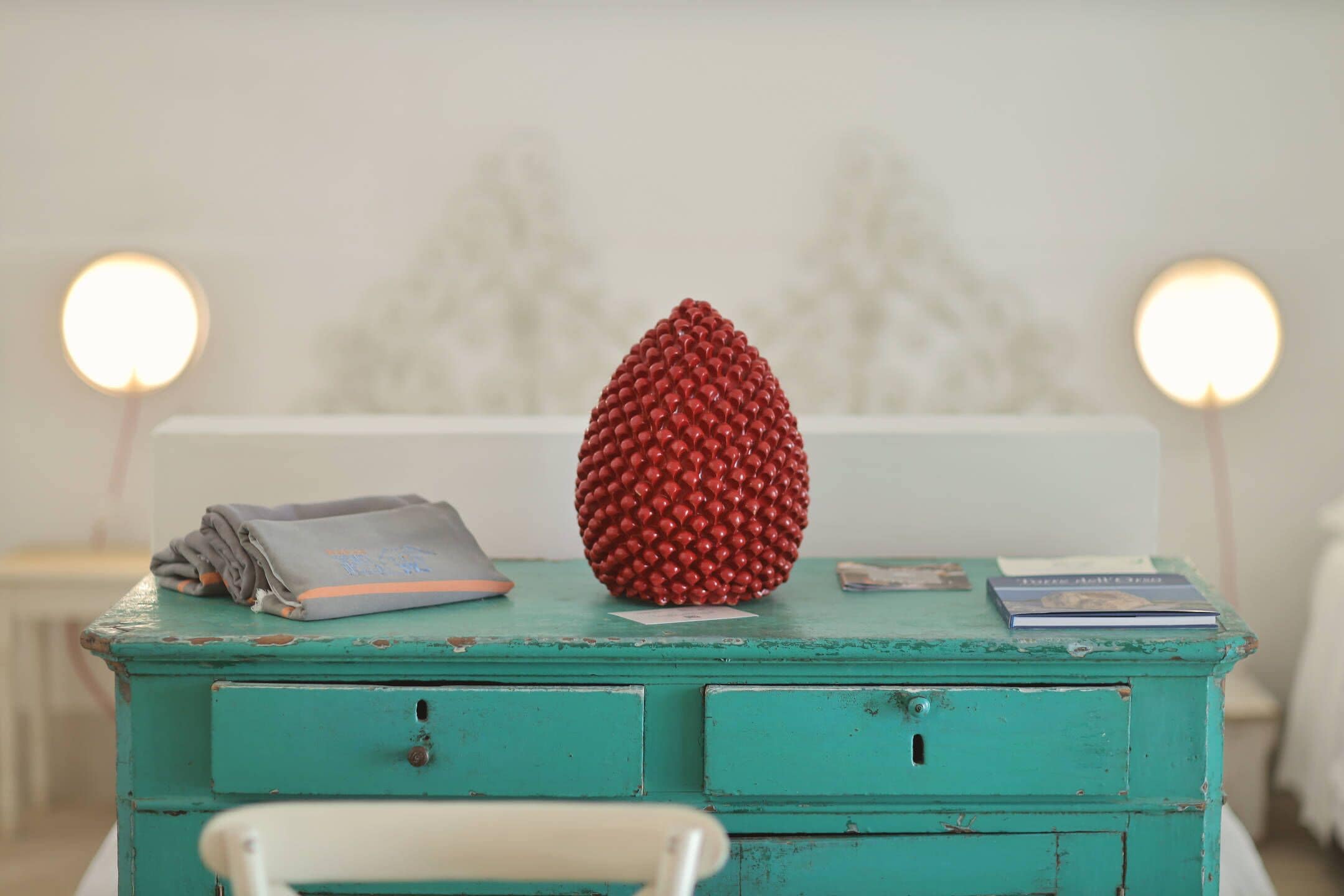 Une commode bleu-vert avec des tiroirs, un objet rouge à la texture particulière posé sur le dessus, et un livre.