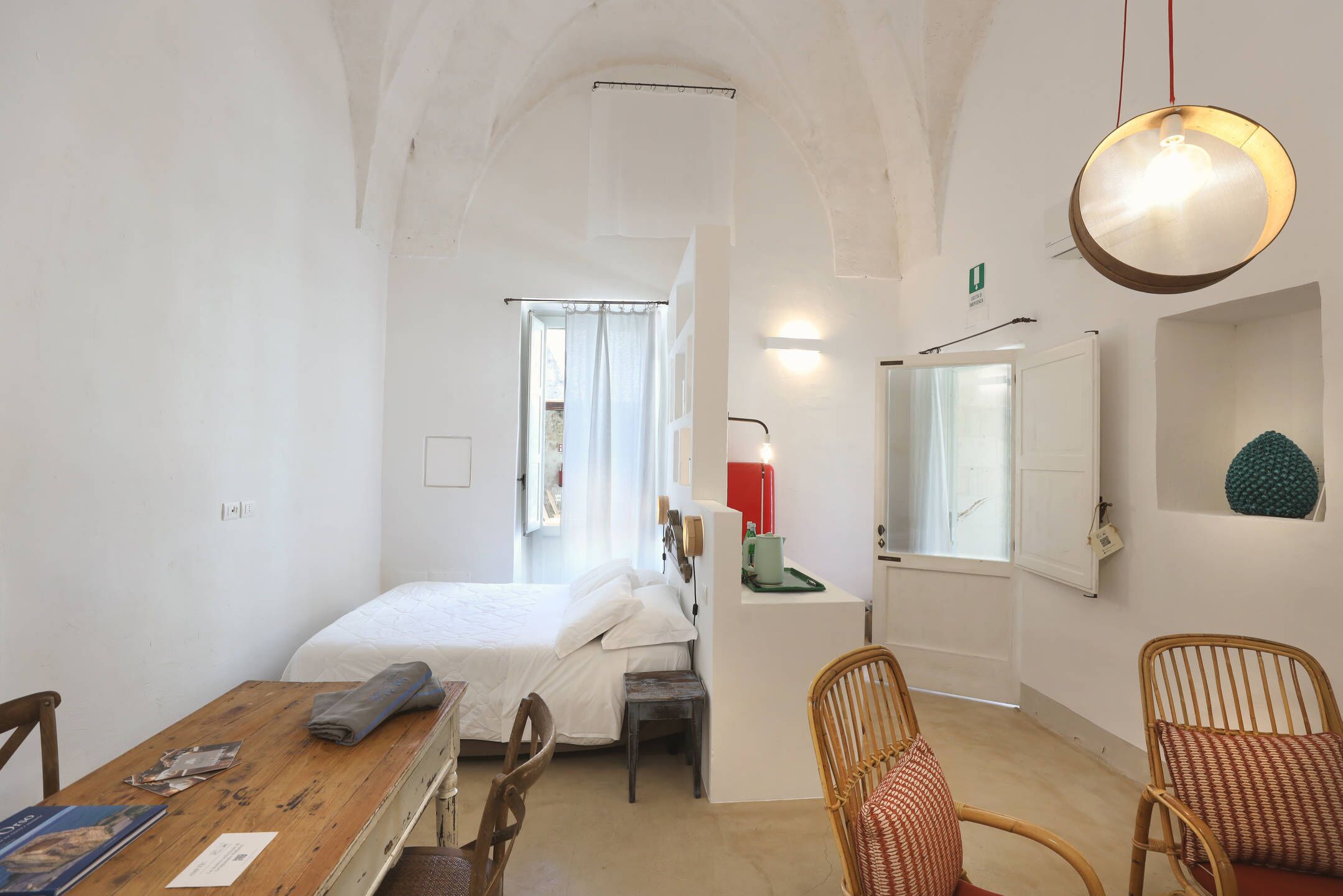 A room with a bed, a wooden table, a chair, a lamp, and a mirror.