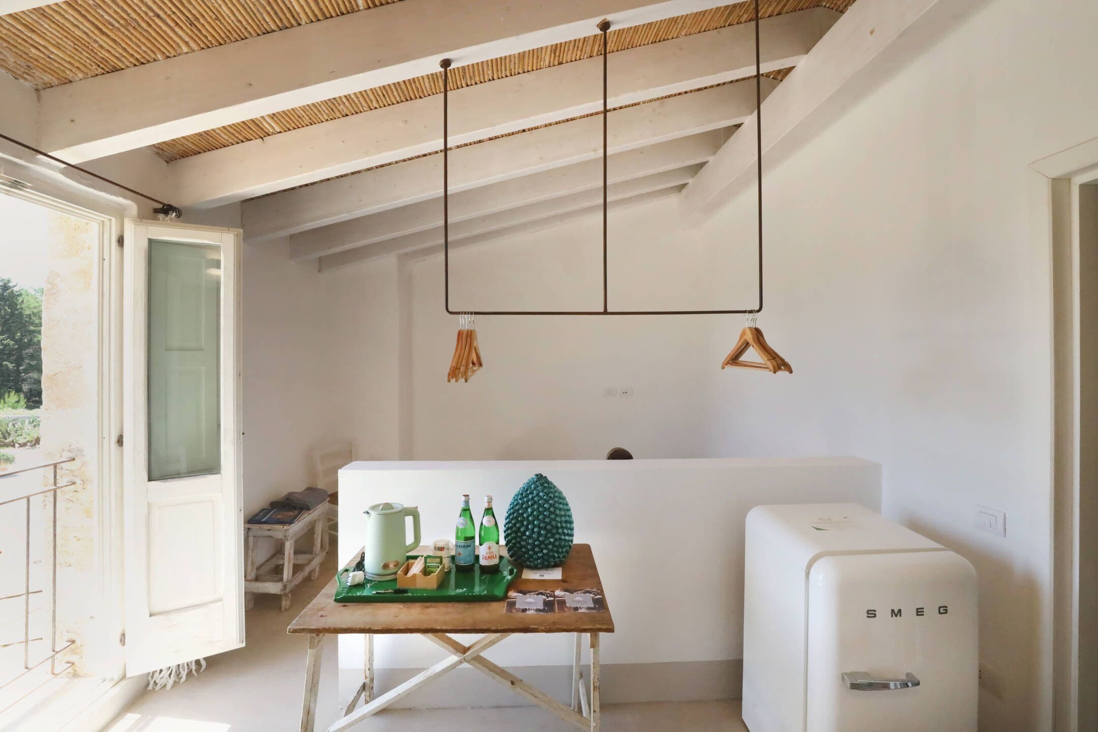 Une cuisine au style minimaliste, avec un réfrigérateur blanc, une table en bois surmontée d’un plateau vert et un luminaire suspendu.