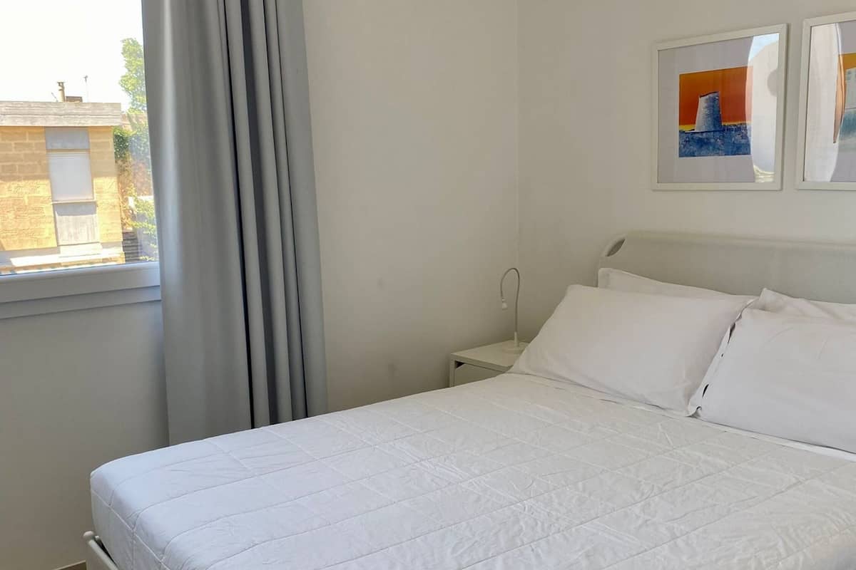 A neatly made bed in a room with a window, a nightstand, and two framed pictures on the wall.