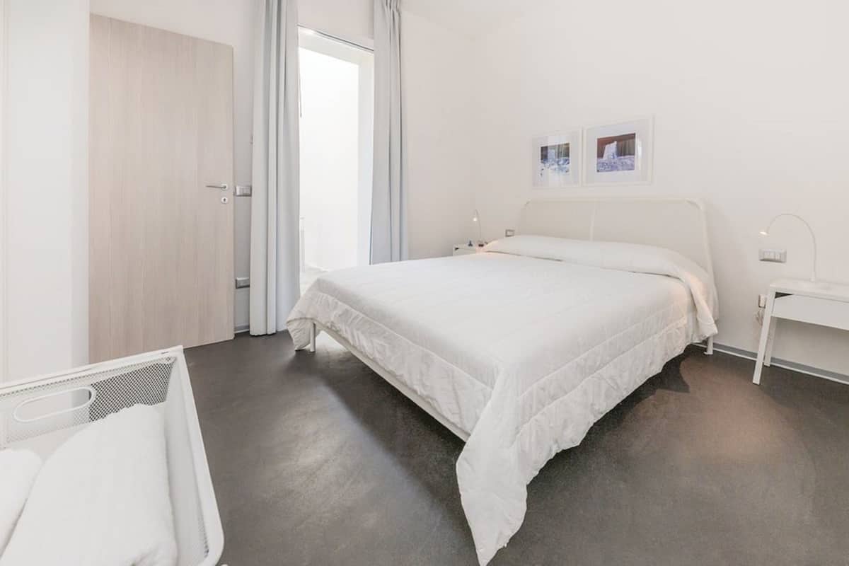A modern hotel room with a large bed, a bedside table, a small desk, and a window with curtains.
