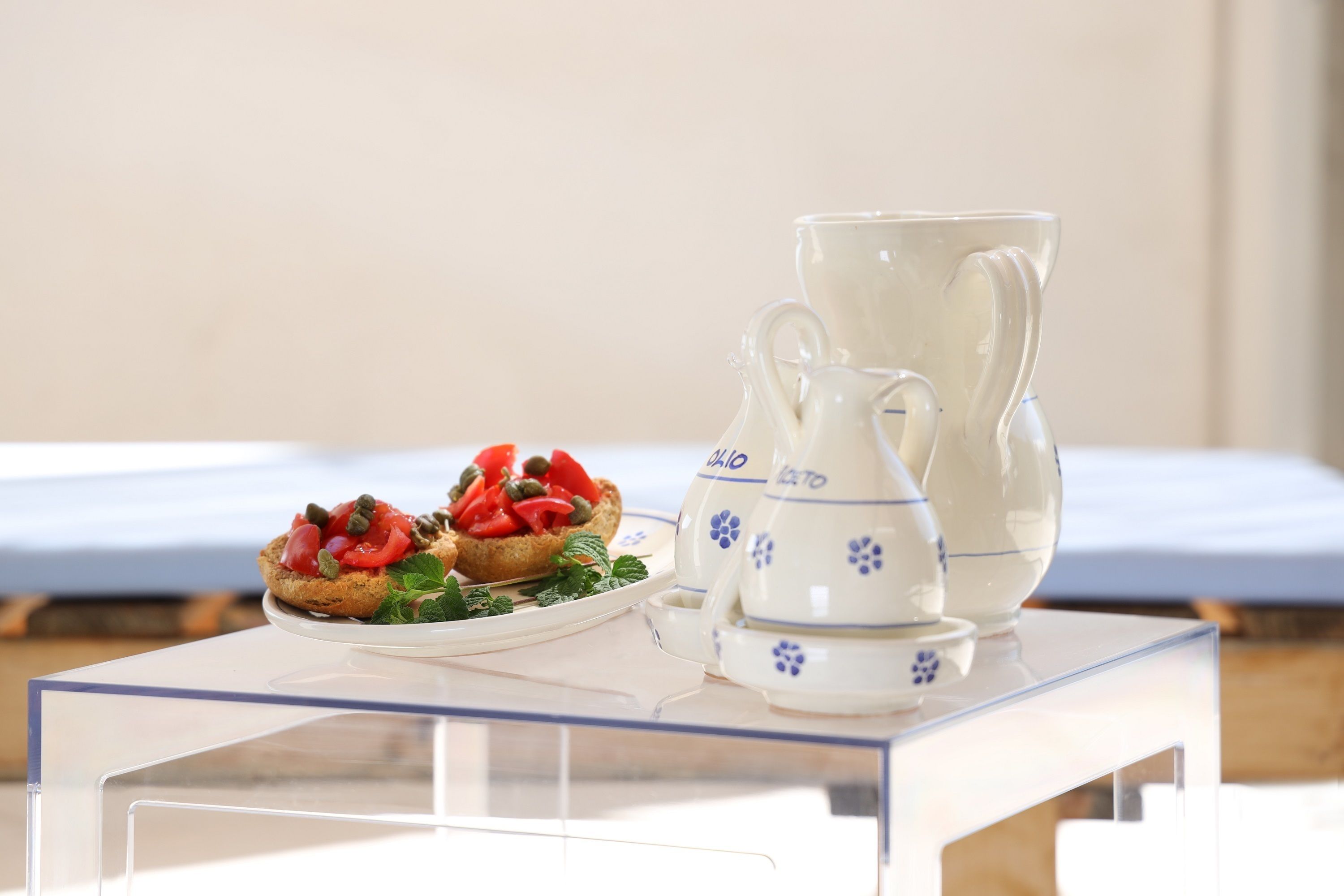 Une table en verre transparent sur laquelle se trouve une assiette de nourriture et deux pichets en céramique blanche ornés de motifs floraux.