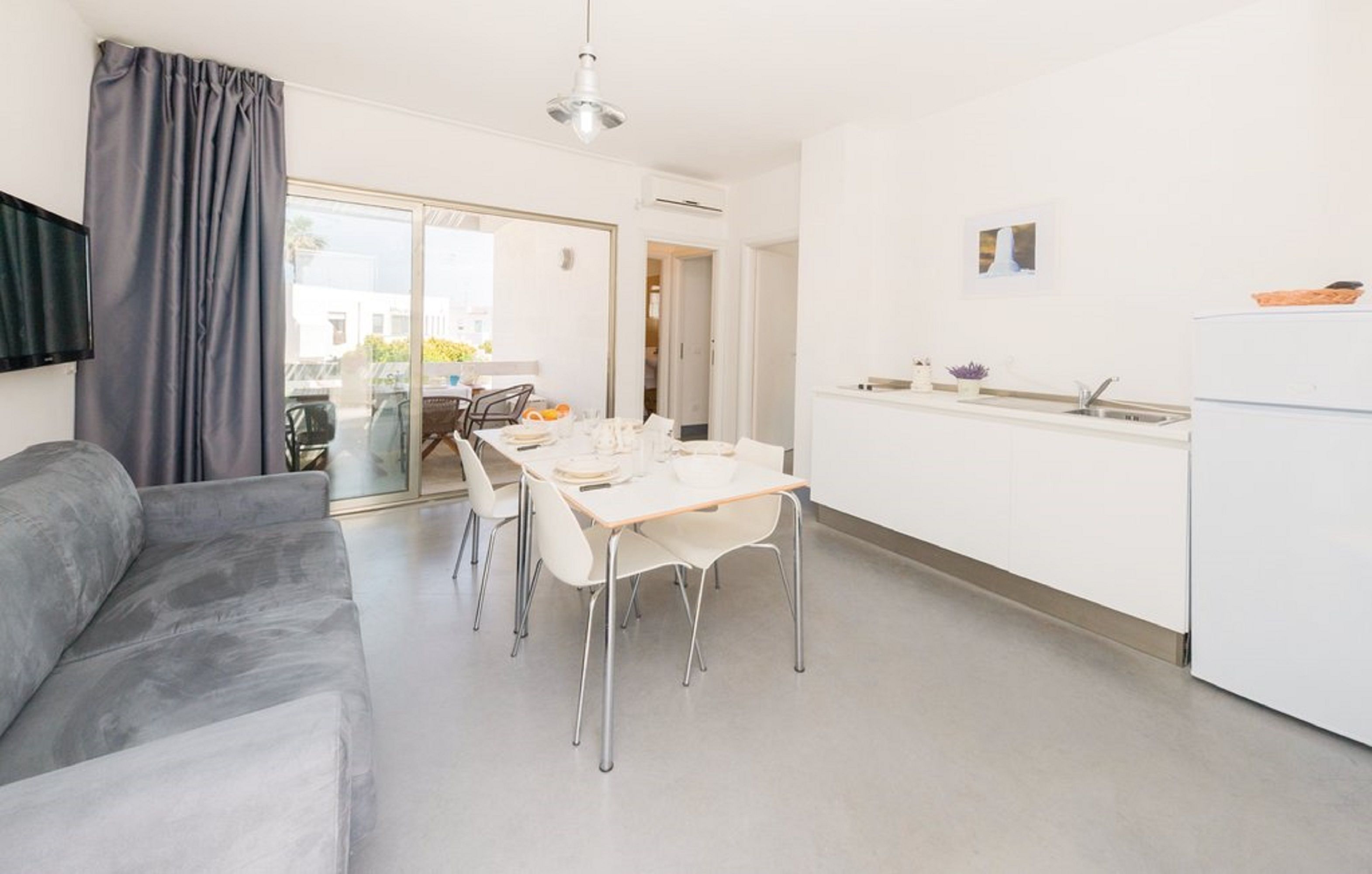 A modern living room with a grey sofa, a dining table with chairs, and a kitchen area with white cabinets and a sink.