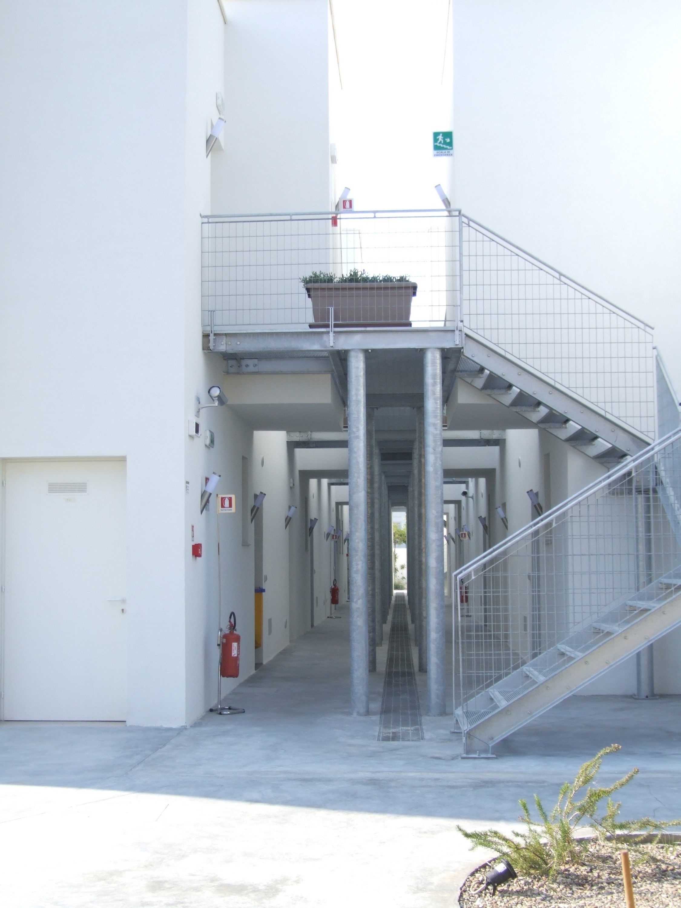 A corridor with a staircase leading to an upper level, fire extinguishers, and a sign indicating an exit.
