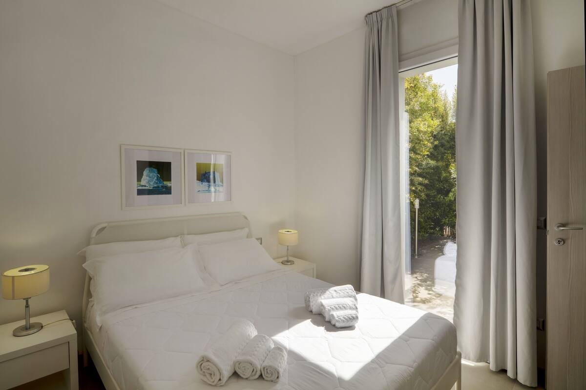 A hotel room with a bed, two bedside lamps, a window with curtains, and a door.