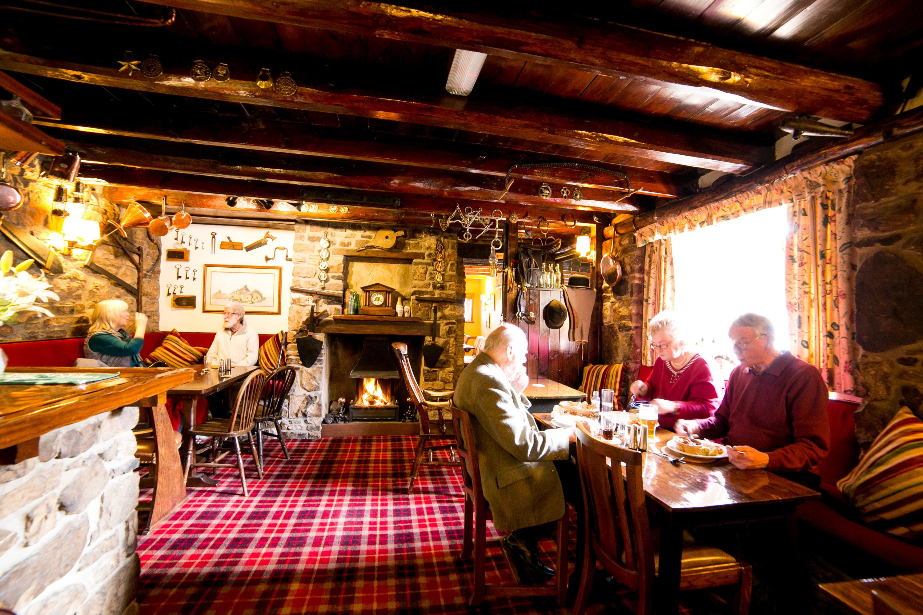 A cozy pub with a stone fireplace, wooden beams, and plaid flooring.