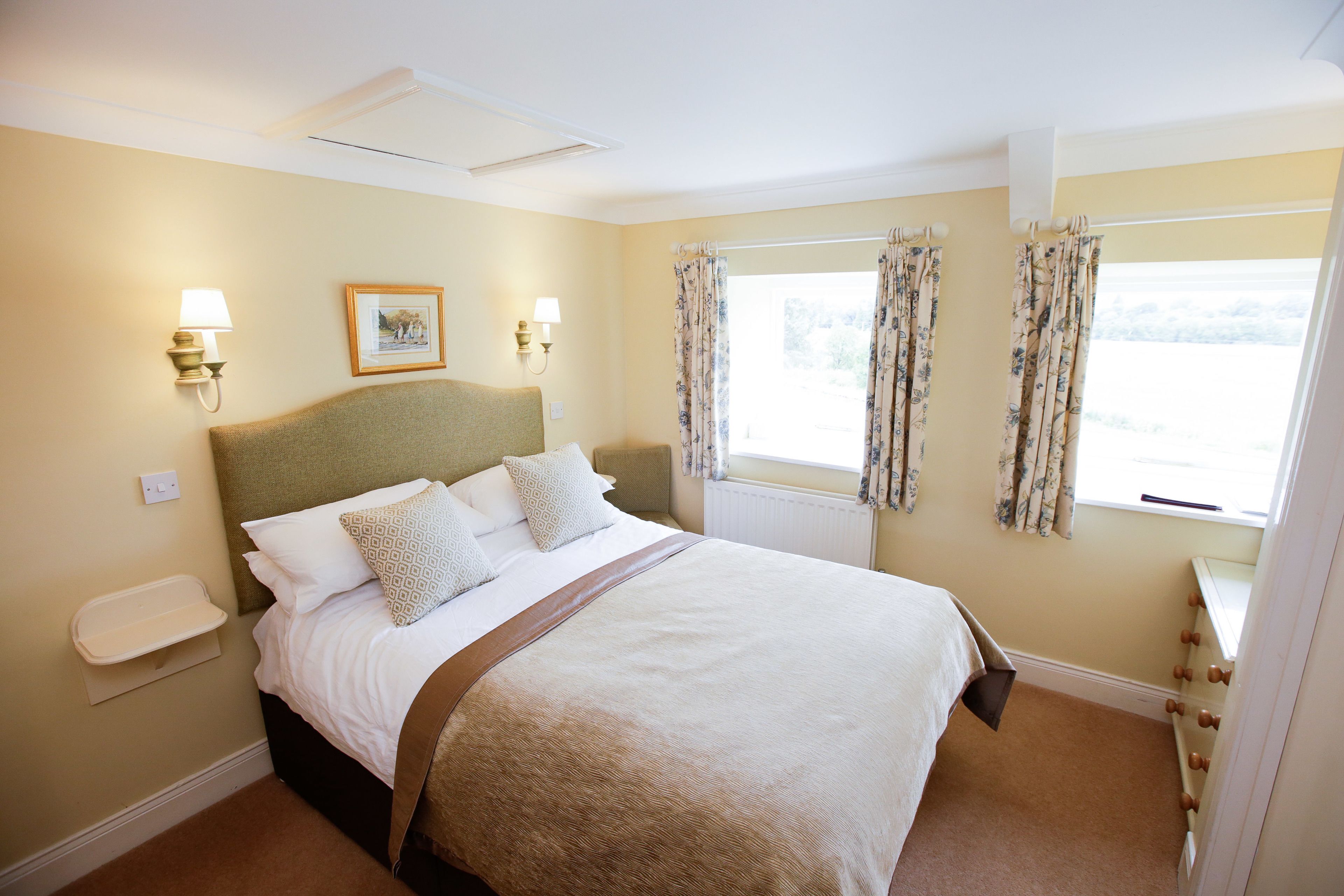 A neatly made bed with a headboard, two bedside lamps, a framed picture on the wall, and a window with floral curtains.