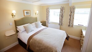 A neatly made bed with a headboard, two bedside lamps, a framed picture on the wall, and a window with floral curtains.