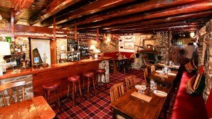 A rustic bar area with wooden beams, a stone wall, and a red plaid floor. There are stools, tables, and a variety of bottles and glasses on display.