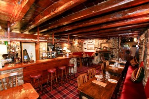 A rustic bar area with wooden beams, a stone wall, and a red plaid floor. There are stools, tables, and a variety of bottles and glasses on display.