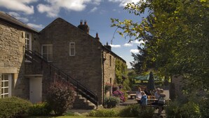 A stone building with a staircase leading to a door, surrounded by well-maintained gardens and a patio area where people are dining.