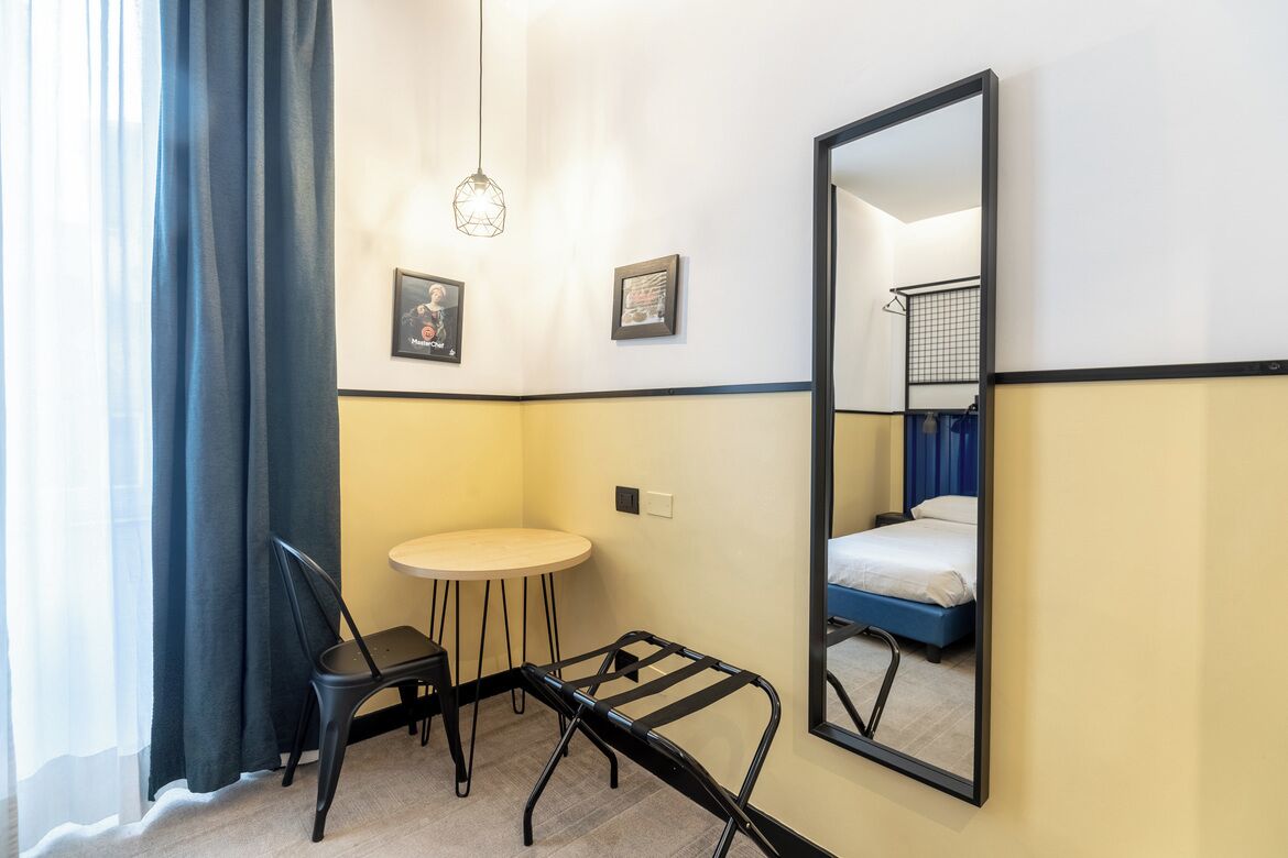 A compact hotel room with a bed, a small round table, a chair, and a mirror on the wall.