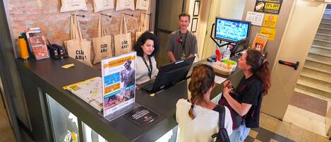 A reception desk with a computer, a poster for a pasta cooking class, and a wall with hanging bags.
