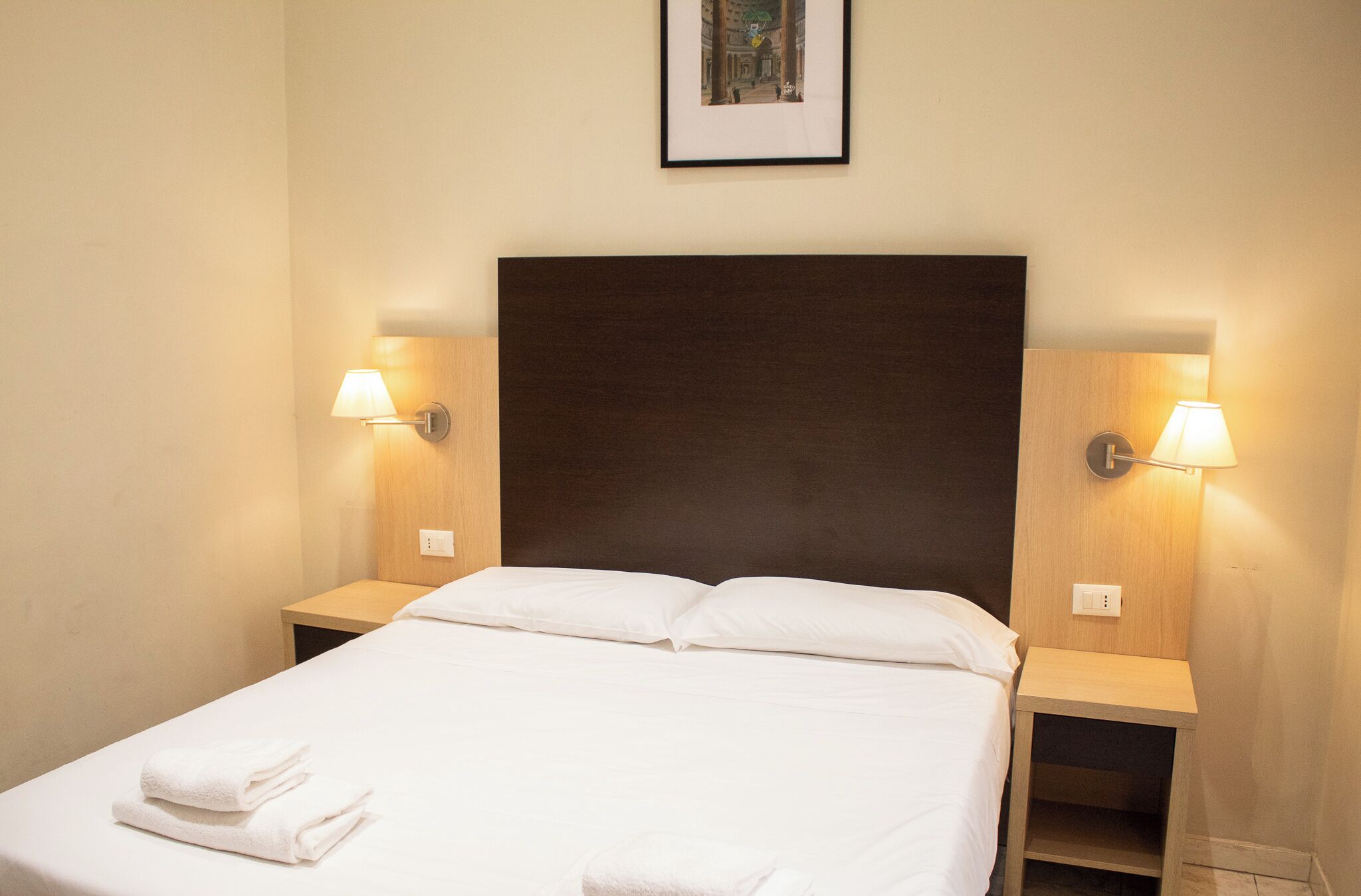A neatly made bed with white linens and two pillows, flanked by two bedside tables with lamps, in a room with a plain wall and a framed picture above the bed.