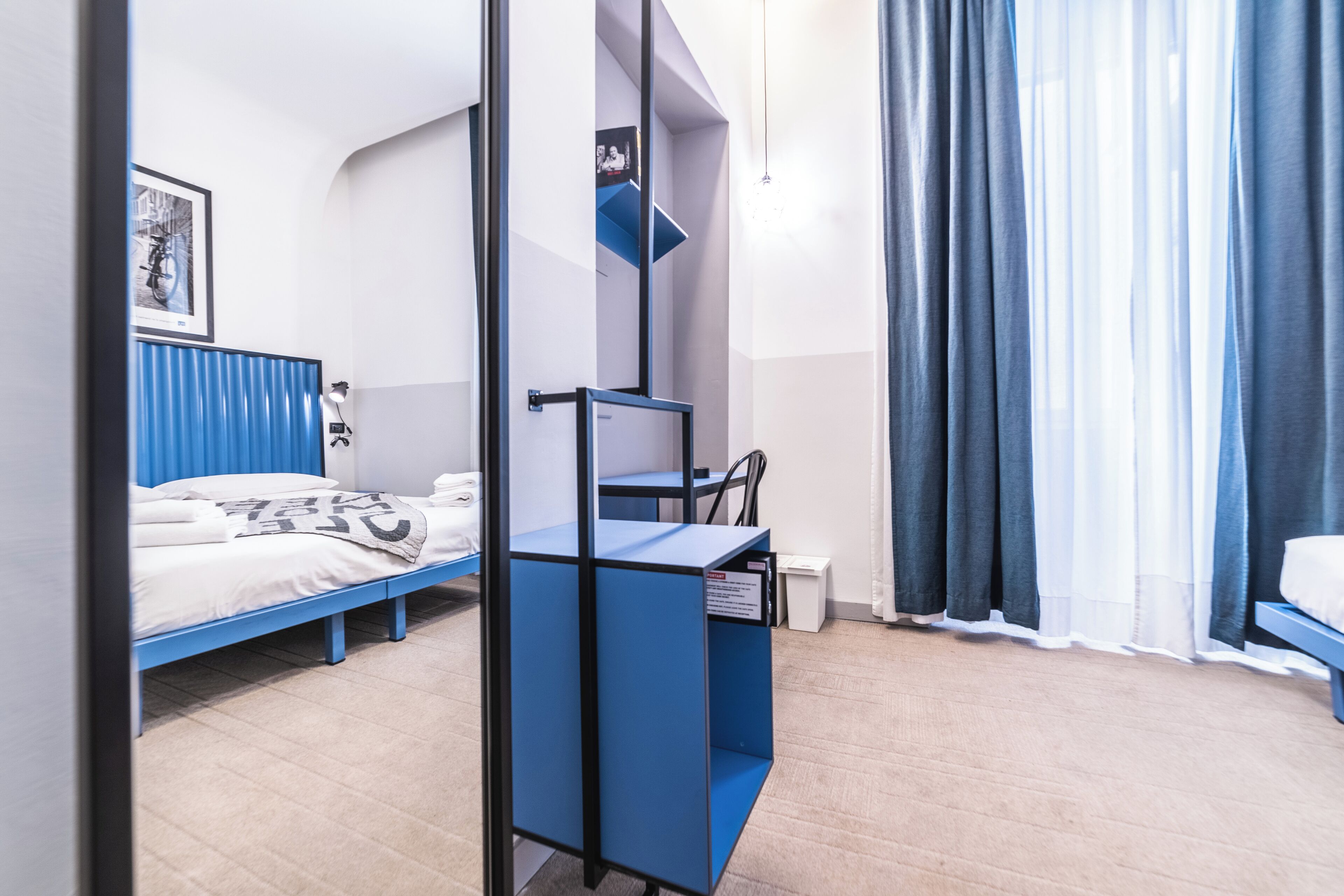 A modern hotel room with a blue bed, a desk with a chair, and a large window with curtains.