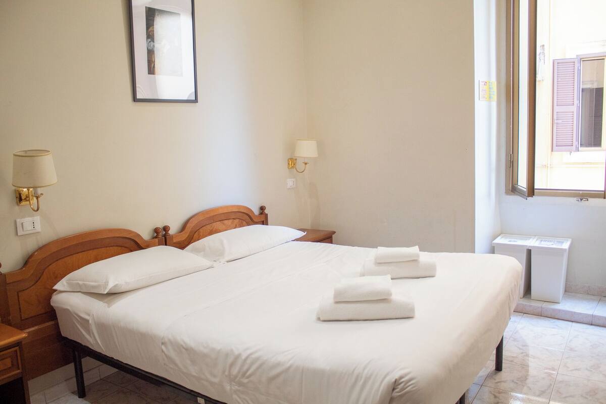 A neatly made bed with white linens and towels, a wooden headboard, a bedside lamp, and a small table beside the bed.