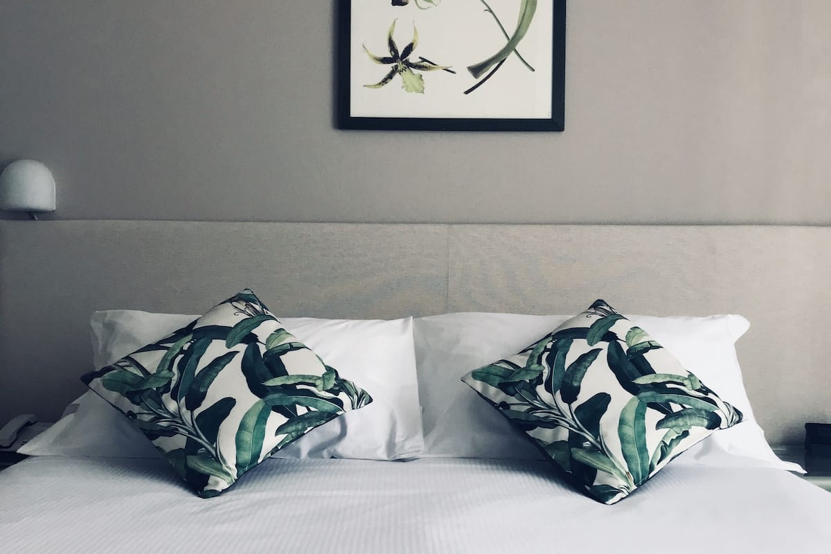 A neatly made bed with white linens and two pillows with a tropical leaf pattern.