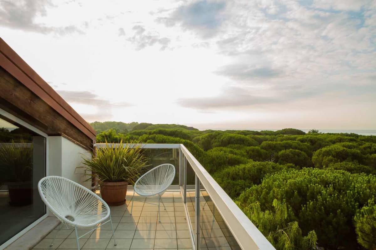 A balcony with two white chairs, a potted plant, and a view of dense greenery.