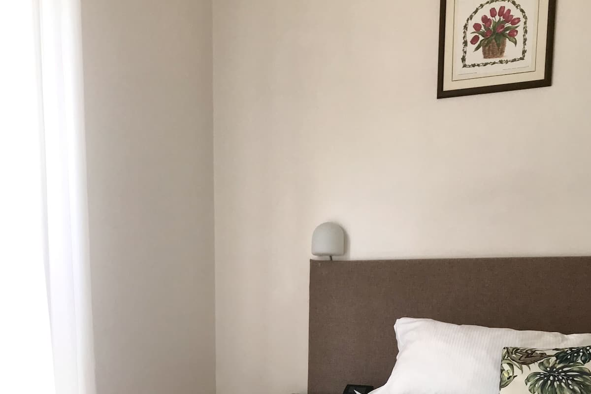 A neatly made bed with white linens, a brown headboard, and a single white pillow. A small bedside table with a lamp is visible to the left.