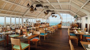 Ein geräumiges Restaurant mit Holzböden und -tischen, großen Fenstern mit Blick auf den Ozean und hängenden Pendelleuchten.