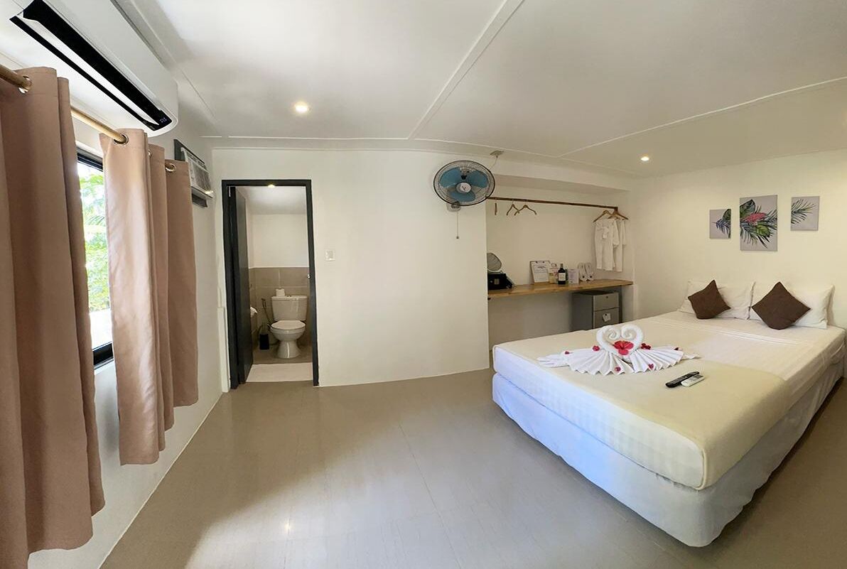 Une chambre d’hôtel comprenant un lit, une table de chevet, un ventilateur, une fenêtre avec des rideaux et une porte donnant sur une salle de bain.