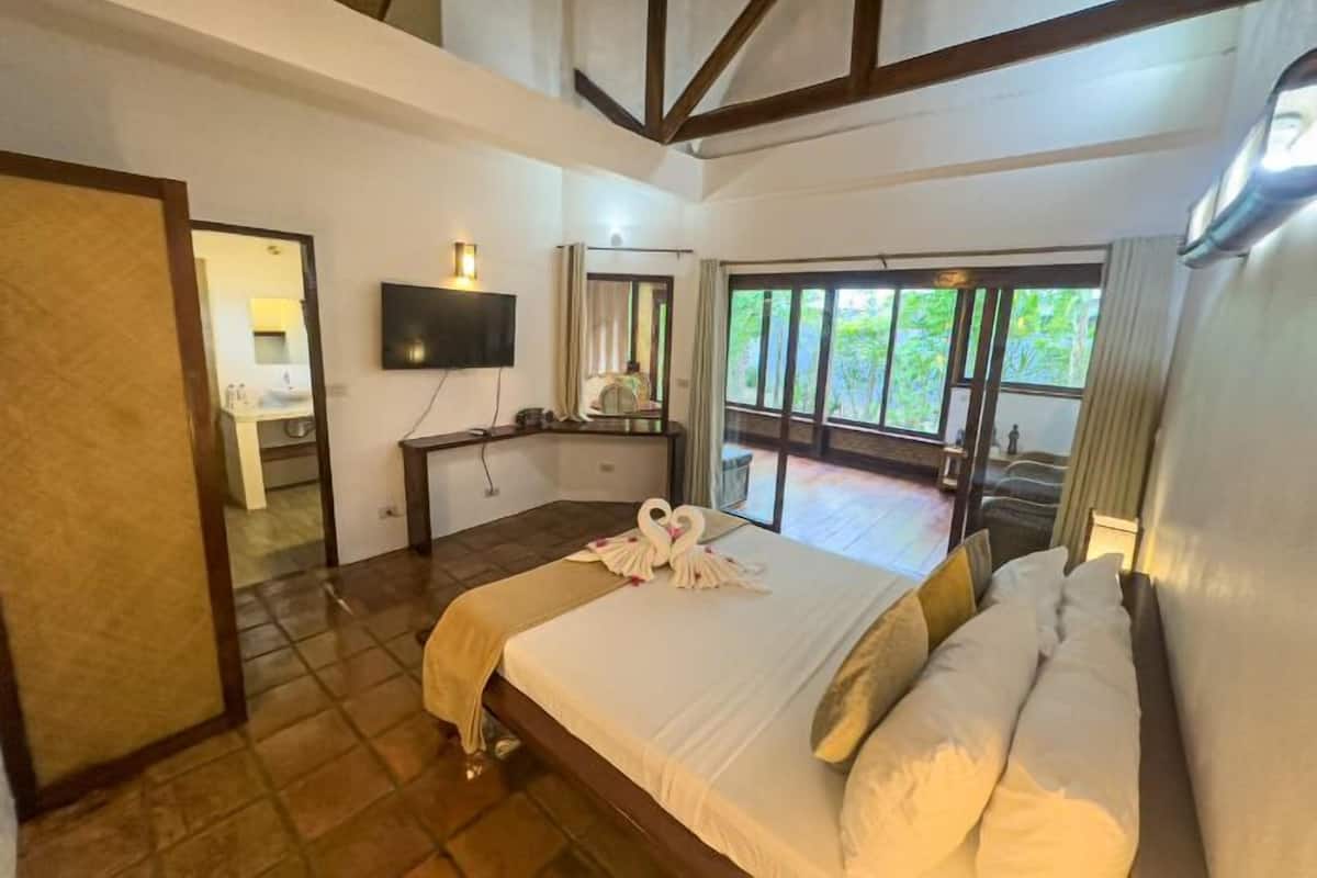 A hotel room with a large bed, a desk, a TV, and a balcony with a view of greenery.