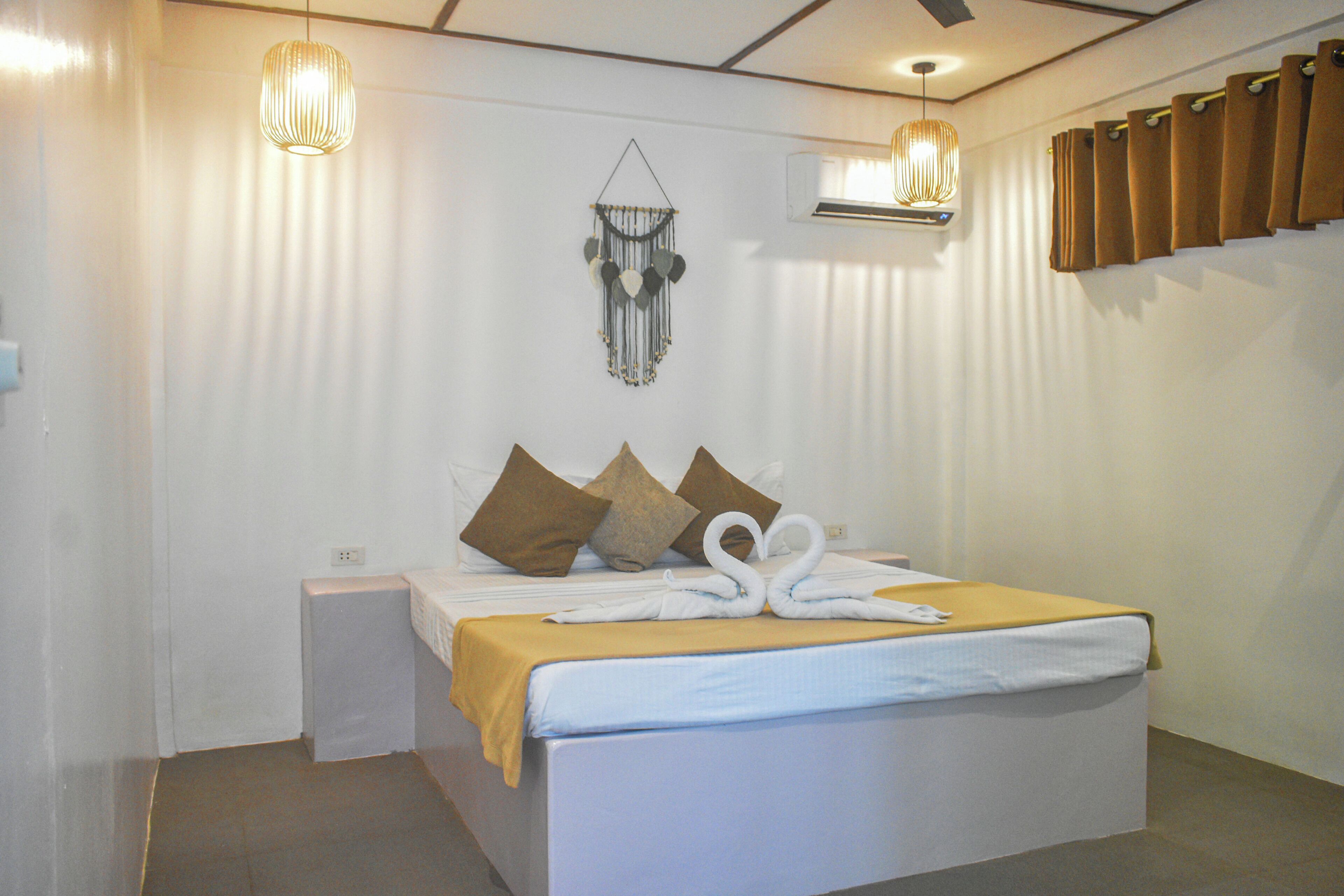 A neatly made bed with a yellow blanket, two white swan-shaped towels, and three brown pillows. There are two pendant lights hanging above the bed and an air conditioning unit on the wall.