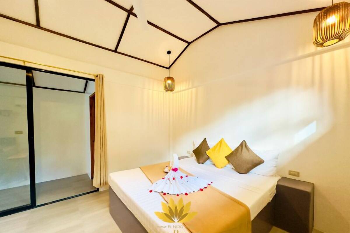 A hotel room with a bed, a bedside table, a window with curtains, and a ceiling light fixture.