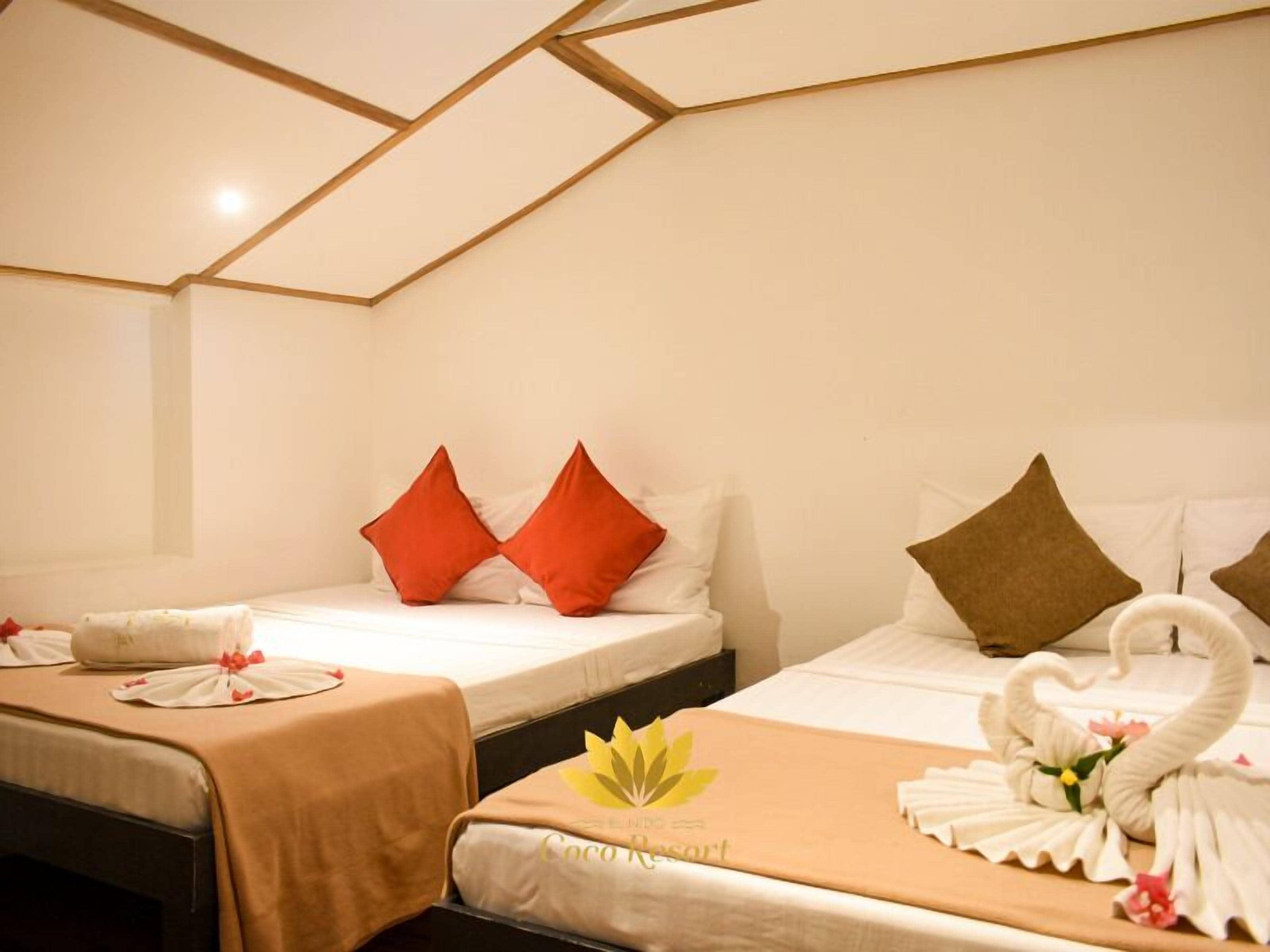 A hotel room with two beds, each with a different pillow color, a swan decoration on one bed, and a bedspread with a logo.
