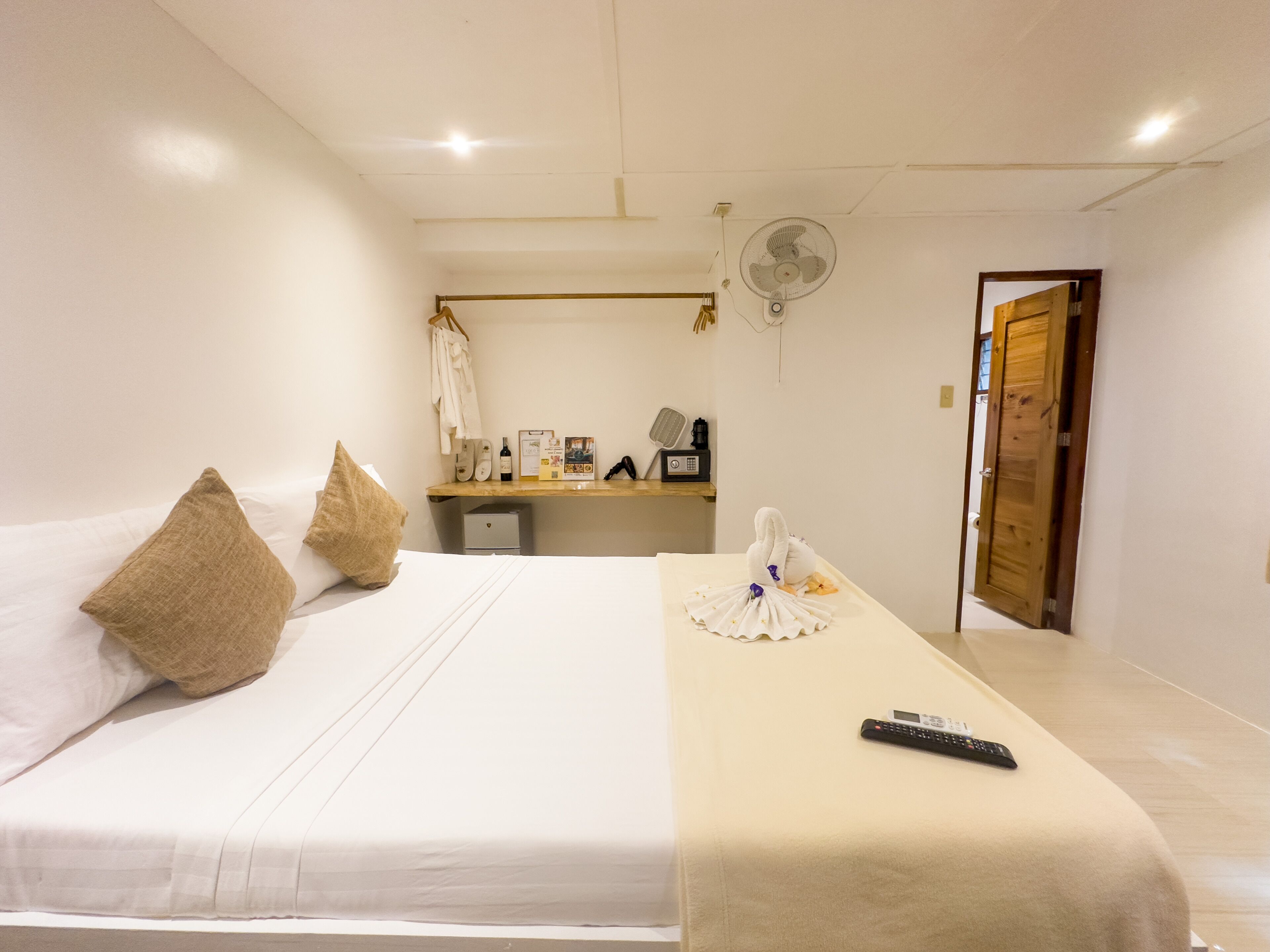A hotel room with a bed, pillows, a bedside table with a remote control, a fan, and a wooden door.