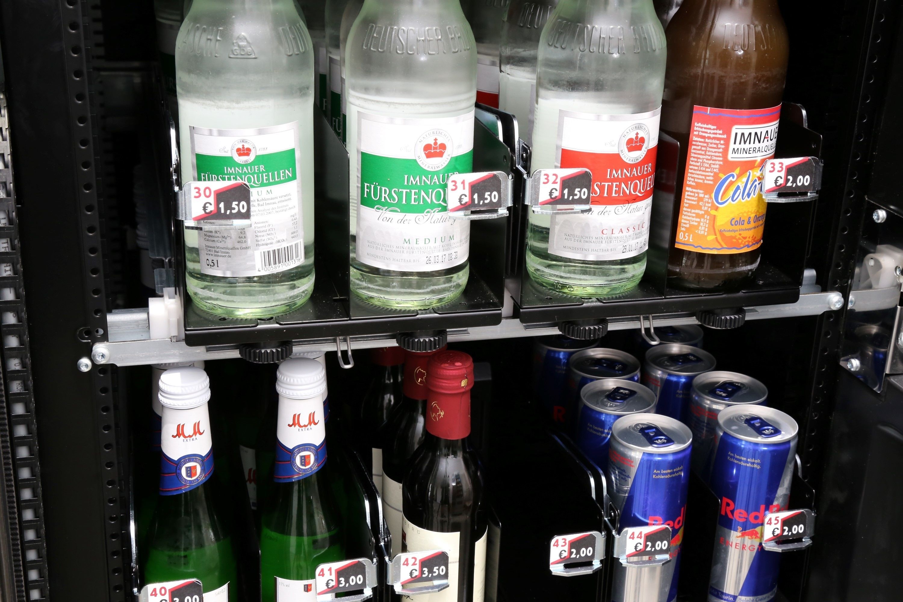 Dispensador de bebidas com diversas opções, incluindo FÜRSTENO, IMNAUER e Cola, além de latas de Red Bull.