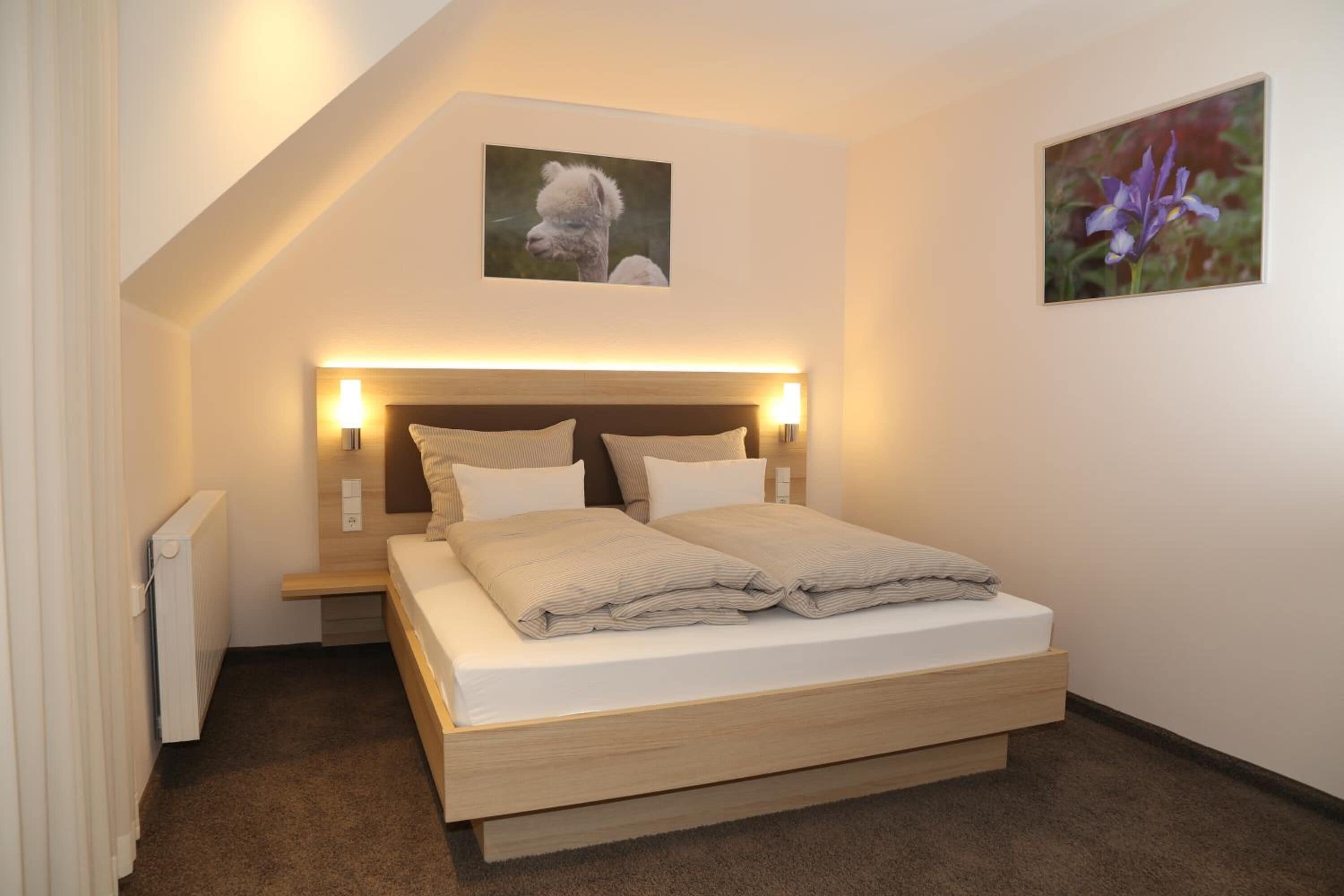 A bedroom with a bed, bedside lamps, a radiator, and two framed pictures on the wall.