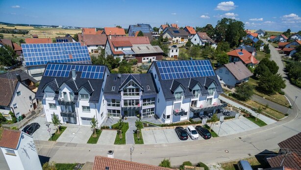 Un edificio residencial con varios paneles solares en el techo, una zona de estacionamiento y vista a la calle.