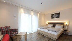 Una habitación de hotel moderna con una cama, un sillón rojo, una mesita y un ventanal con cortinas translúcidas.
