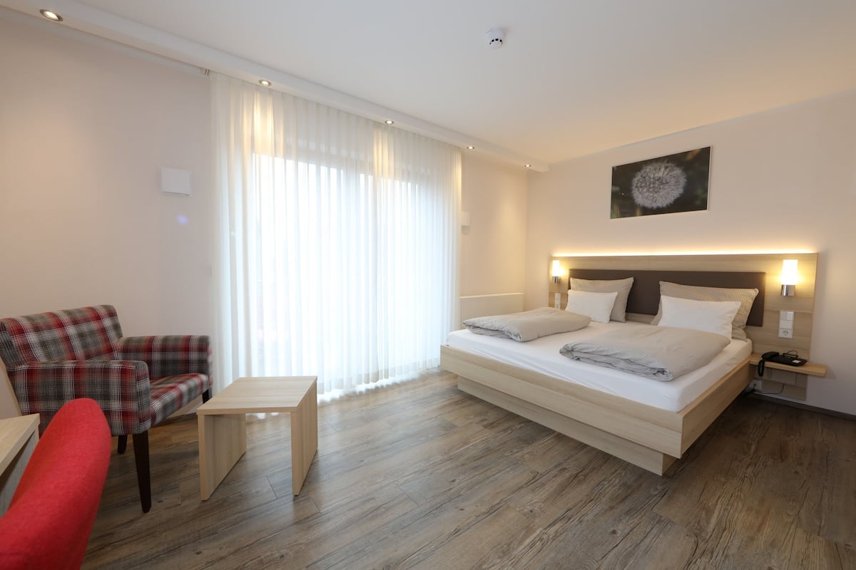A modern hotel room with a bed, a red armchair, a small table, and a large window with sheer curtains.