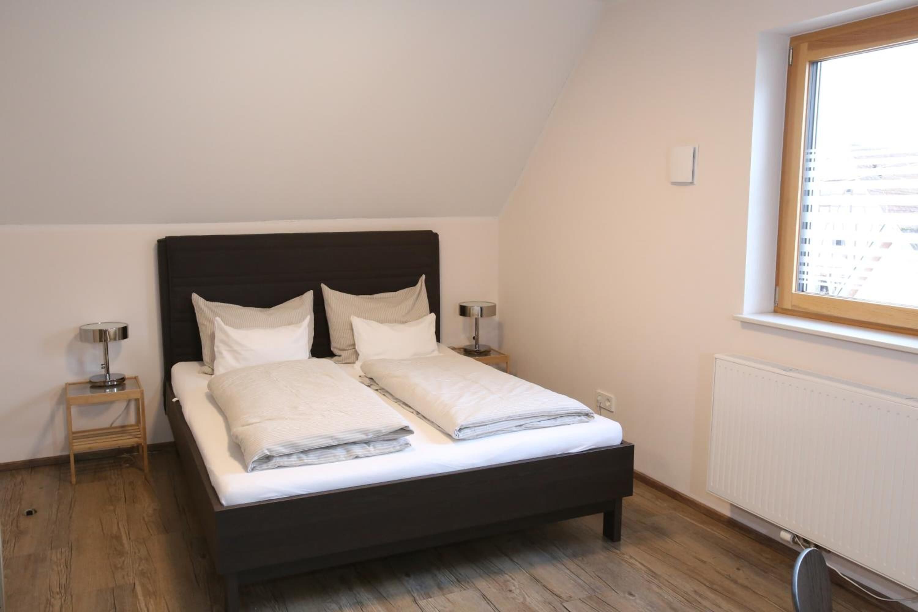 A bedroom with a dark headboard, two bedside tables with lamps, a window with a view of buildings, and a radiator.