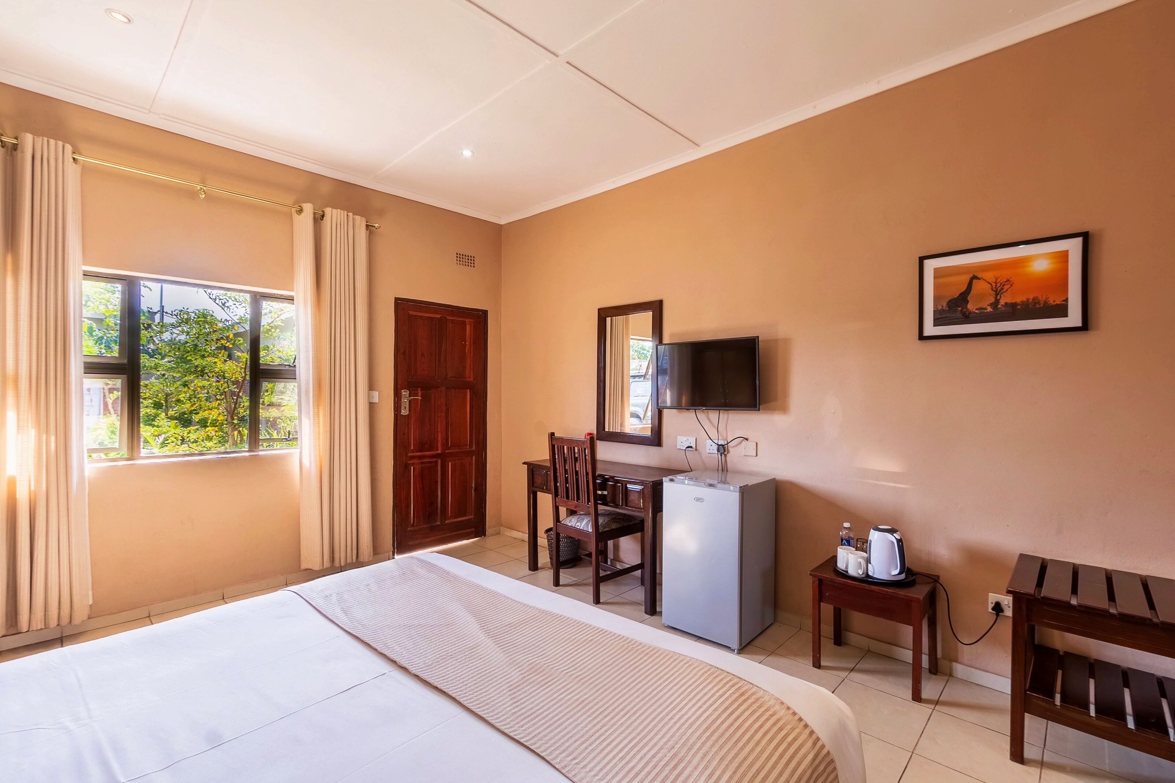 Habitación de hotel con cama, escritorio con silla, televisor, mesita con hervidor y un cuadro en la pared.